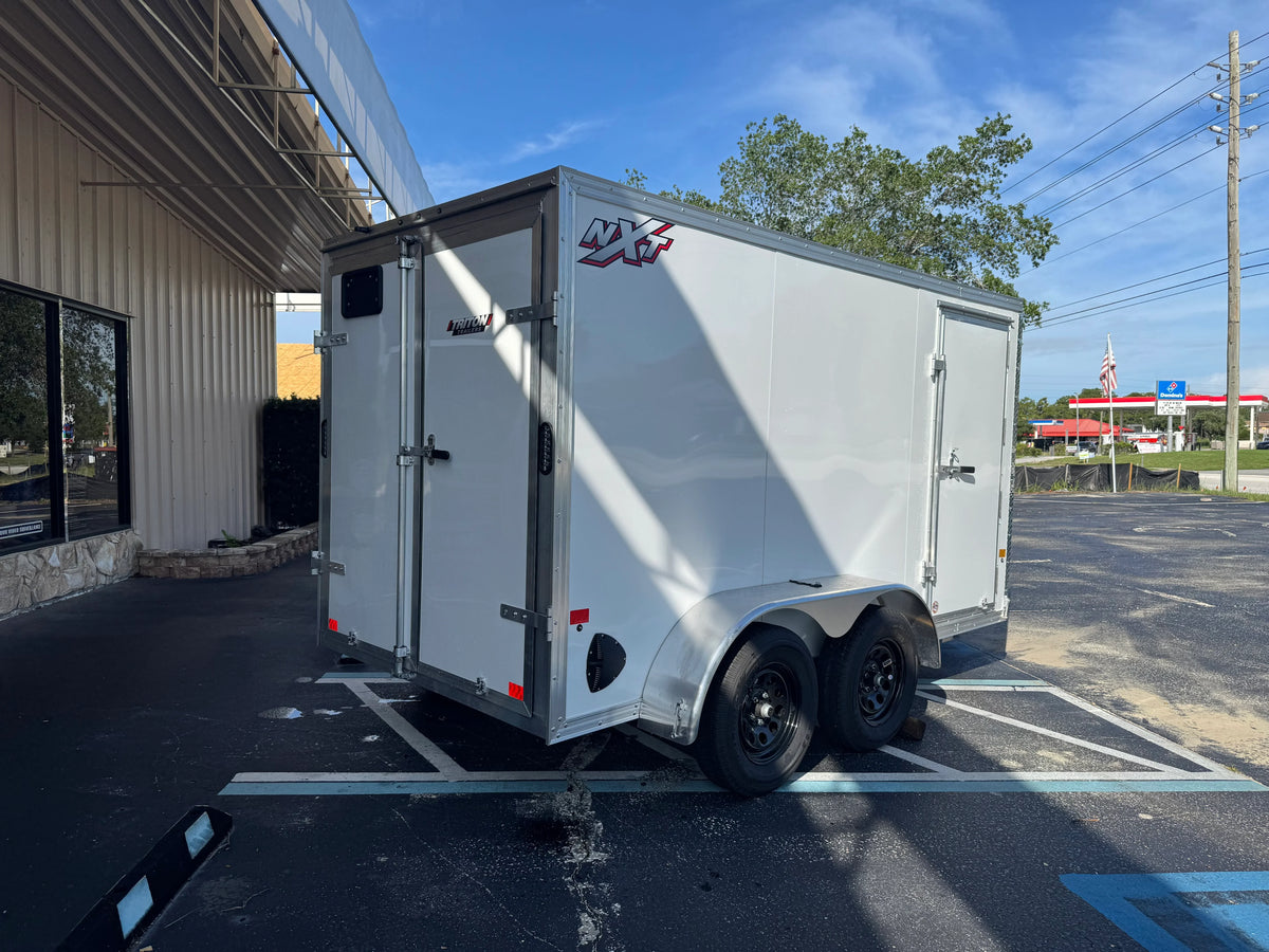 White enclosed cargo trailer with NXT and Triton Trailers branding, parked outside a commercial building.